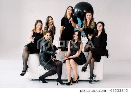 Cheerful young female friends in elegant black dresses with martini drinks enjoying birthday party or New Year celebration on white sofa at light room Cheerful young female friends in elegant black dresses with martini drinks enjoying birthday party or New Year celebration on white sofa at light room 65533845