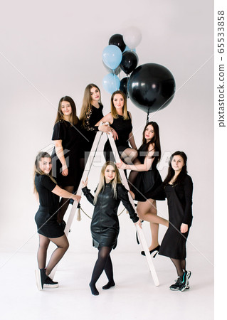 Happy young seven girls in black dresses having fun celebrating birthday in light room. The friends having fun posing on the ladder, decorated with colorful air balloons. Happy young seven girls in black dresses having fun celebrating birthday in light room. The friends having fun posing on the ladder, decorated with colorful air balloons. 65533858