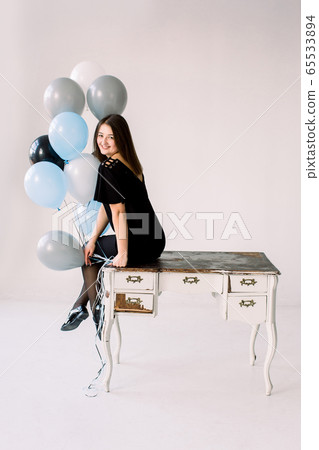 Happy emotional glamour smiling young woman in black dress and shoes with birthday holiday ballons sitting on white wooden vintage table indoor 65533894