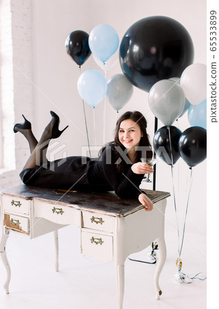 Attractive young girl in black dress lying on white vintage table, holding the martini glass, on white background. Pretty girl elebrating Happy New Year, birthday party balloons on the background Attractive young girl in black dress lying on white vintage table, holding the martini glass, on white background. Pretty girl elebrating Happy New Year, birthday party balloons on the background 65533899