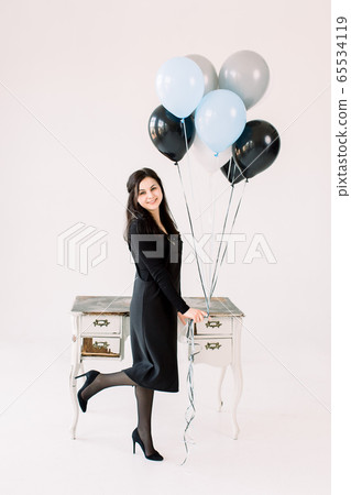 glad positive Caucasian girl in black dress holds bunch of air balloons, standing near vintage white table with drawers, waits for guests, isolated over white background. Birthday party concept glad positive Caucasian girl in black dress holds bunch of air balloons, standing near vintage white table with drawers, waits for guests, isolated over white background. Birthday party concept 65534119