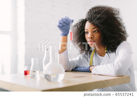 Young beautiful African American girl doctor in a white coat with a stethoscope. sitting at a table with reagent flasks on white background. Investigator checking test tubes. Young beautiful African American girl doctor in a white coat with a stethoscope. sitting at a table with reagent flasks on white background. Investigator checking test tubes. 65537241