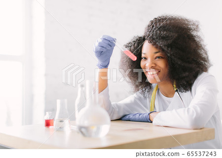 Young beautiful African American girl doctor in a white coat with a stethoscope. sitting at a table with reagent flasks on white background. Female lab assistant studying blood sample for analysis Young beautiful African American girl doctor in a white coat with a stethoscope. sitting at a table with reagent flasks on white background. Female lab assistant studying blood sample for analysis 65537243