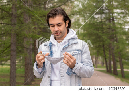 Young contemporary man in hoodie and denim jacket holding protective mask Young contemporary man in hoodie and denim jacket holding protective mask 65542891