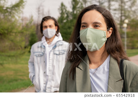 Pretty young brunette woman in protective mask walking in public park 65542918