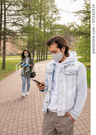 Serious young man in casualwear standing in public park and scrolling in phone 65542919