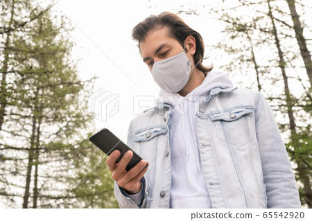 Young man in denim jacket, hoodie and mask scrolling or texting in smartphone 65542920