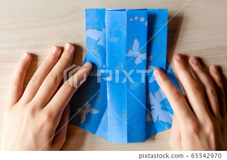Girl's hands making origami blue dress from butterfly printed paper. Step by step instruction, step13. Mother's day, birhday, summer gift, family leisure time concept. DIY tutorial, flatlay, top view 65542970