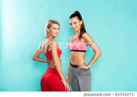 Girls power. Two young and attractive sporty girls in sportswear looking at camera and smiling while standing against blue background. Sport and fitness Girls power. Two young and attractive sporty girls in sportswear looking at camera and smiling while standing against blue background. Sport and fitness 65543271