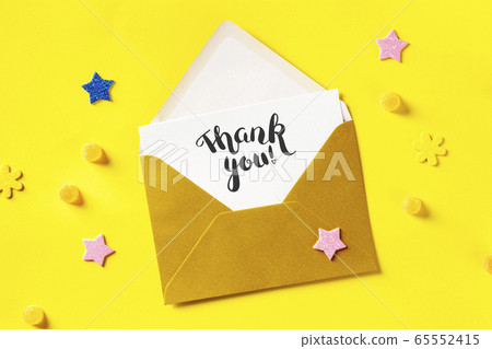 Thank you card in a golden envelope, shot from the top on a yellow background with glitter stars 65552415