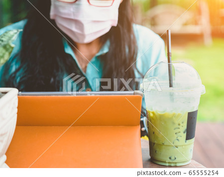 An ice green tea for woman wearing protective mask and working from home with tablet during Coronavirus or Covid-19 quarantine. Coronavirus outbreak and flu covid epidemic. Selective focus on straw. An ice green tea for woman wearing protective mask and working from home with tablet during Coronavirus or Covid-19 quarantine. Coronavirus outbreak and flu covid epidemic. Selective focus on straw. 65555254