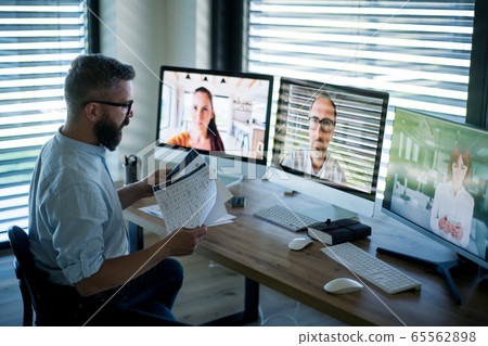 Side view of businessman sitting at desk, having video call. Side view of businessman sitting at desk, having video call. 65562898