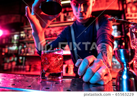 Close up of barman finishes preparation of alcoholic cocktail, pouring drink in multicolored neon light, focus on glass Close up of barman finishes preparation of alcoholic cocktail, pouring drink in multicolored neon light, focus on glass 65563229