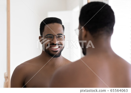 Smiling biracial man in glasses look in mirror in bathroom Smiling biracial man in glasses look in mirror in bathroom 65564063