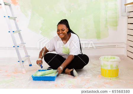 Pretty smiling african american woman painting interior wall of home with paint roller. Redecoration, renovation, apartment repair and refreshment concept. 65568380