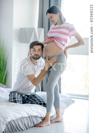 Bearded man putting his ear close to his pregnant wifes abdomen and looking excited Bearded man putting his ear close to his pregnant wifes abdomen and looking excited 65569155