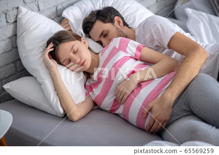 Dark-haired bearded man hugging his pregnant wife while sleeping and looking peaceful Dark-haired bearded man hugging his pregnant wife while sleeping and looking peaceful 65569259