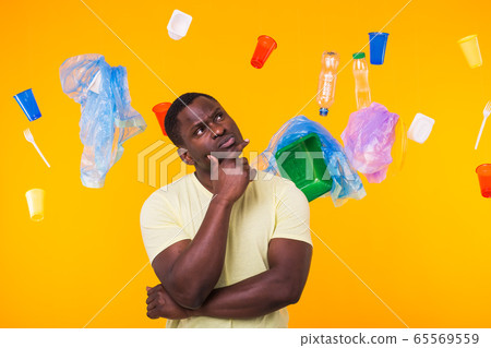 Problem of trash, plastic recycling, pollution and environmental concept - Serious african american man looking on trash on yellow background. He is thinking about ecology 65569559