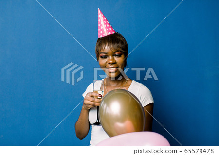 Celebration Birthday Concept - Close up Portrait happy young beautiful african woman in white t-shirt and pink party hat smiling with colorful party balloon. Celebration Birthday Concept - Close up Portrait happy young beautiful african woman in white t-shirt and pink party hat smiling with colorful party balloon. 65579478