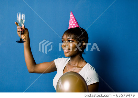 Portrait of beautiful birthday african girl resting at party. Pretty smiling african girl holding glass with champagne and air balloons posing on blue background Portrait of beautiful birthday african girl resting at party. Pretty smiling african girl holding glass with champagne and air balloons posing on blue background 65579479