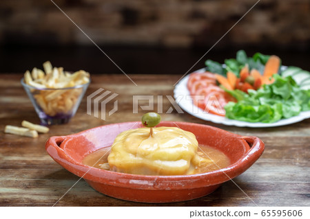 Portuguese francesine in a clay plate with salad and fried potatoes on a wooden background. Portuguese francesine in a clay plate with salad and fried potatoes on a wooden background. 65595606