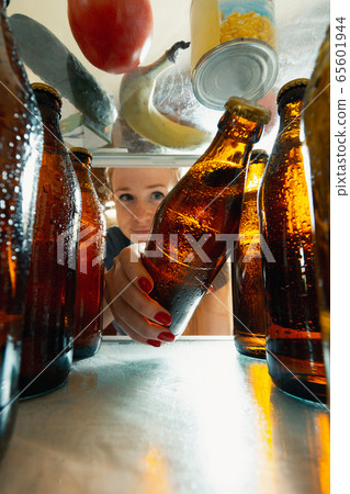 Caucasian woman takes cold refreshing beer from out the fridge, inside view from fridge of hand holding the bottle Caucasian woman takes cold refreshing beer from out the fridge, inside view from fridge of hand holding the bottle 65601944
