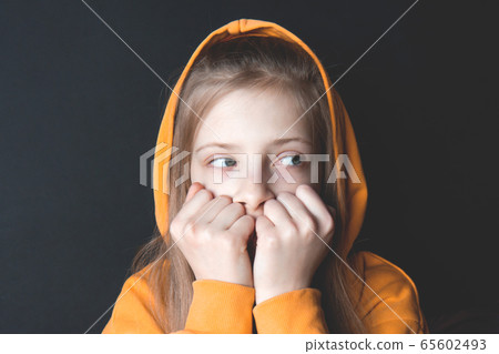 Teenager girl in an orange sweatshirt with bright emotions on her face on a black background. Covered her mouth with hands. Selective focus. Side view 65602493