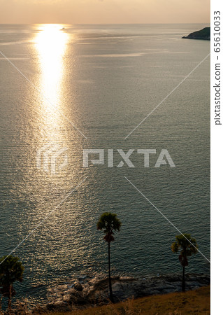 View on Palm tree and islands near sea water during beautiful sunset with a bright yellow sun shining rays over the rocks. Suited for a wallpaper background with a soft yellow sky. Sun over water. 65610033
