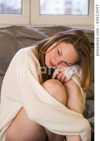 Woman sitting on bed in sweater hugging her knees Woman sitting on bed in sweater hugging her knees 65612047