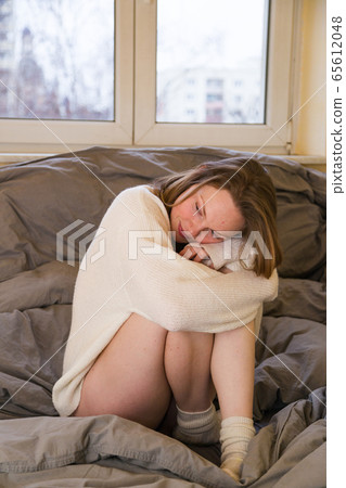 Woman sitting on bed in sweater hugging her knees Woman sitting on bed in sweater hugging her knees 65612048