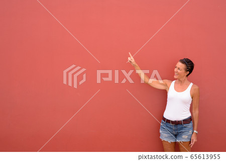 Happy mature beautiful tourist woman pointing up against pink wall Happy mature beautiful tourist woman pointing up against pink wall 65613955
