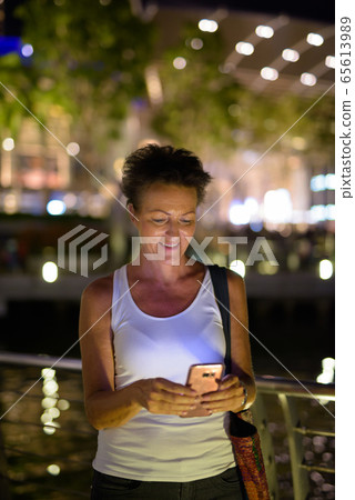 Happy mature beautiful tourist woman using phone in the city at night Happy mature beautiful tourist woman using phone in the city at night 65613989