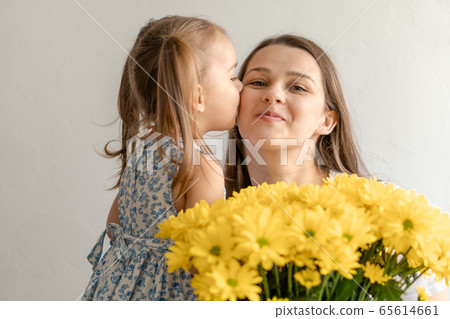motherhood, mothers day, birthday, childhood, family concept - close-up little girl in dress congratulate and give young mother bouquet of yellow daisy flowers and kisses her on white background. 65614661
