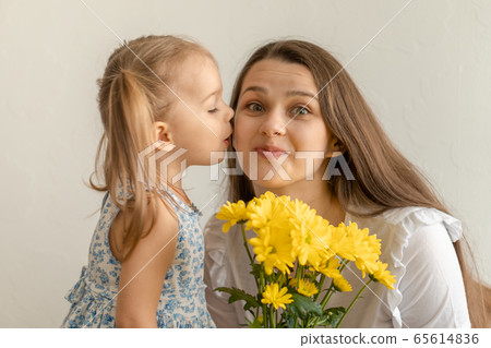 motherhood, mothers day, birthday, childhood, family concept - close-up little girl in dress congratulate and give young mother bouquet of yellow daisy flowers and kisses her on white background. 65614836