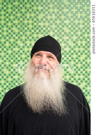 Mature bearded hipster man with beanie hat thinking against green wall 65616351