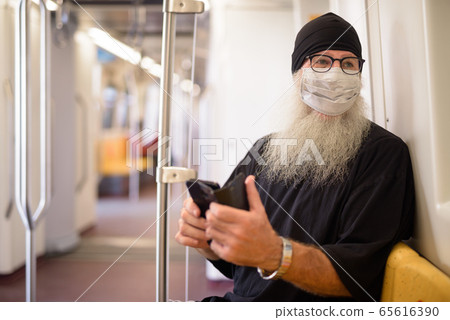 Mature bearded hipster man with mask thinking while using phone inside the train 65616390