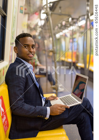 Happy young handsome African businessman using laptop while sitting with distance inside the train 65616448