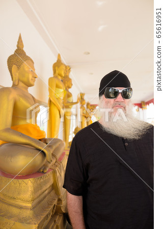 Mature bearded tourist man with sunglasses beside golden Buddha statue at the temple 65616951