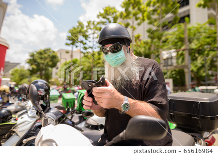 Mature bearded hipster man with sunglasses and mask using phone while sitting on motorcycle 65616984