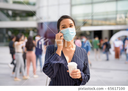 Young Asian businesswoman with mask talking on the phone while having coffee on the go as the new Young Asian businesswoman with mask talking on the phone while having coffee on the go as the new 65617100
