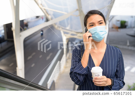 Young Asian businesswoman with mask talking on the phone while having coffee on the go as the new Young Asian businesswoman with mask talking on the phone while having coffee on the go as the new 65617101