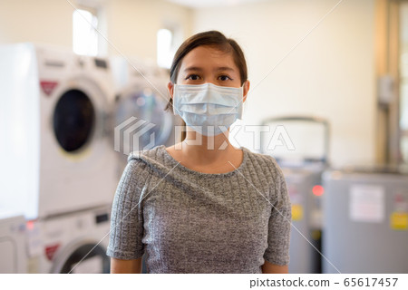 Young Asian woman wearing mask at the laundromat for protection from corona virus outbreak Young Asian woman wearing mask at the laundromat for protection from corona virus outbreak 65617457