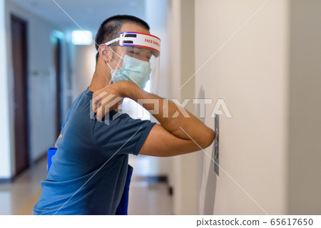 Young Asian man with mask and face shield pressing elevator button with elbow for prevention of Young Asian man with mask and face shield pressing elevator button with elbow for prevention of 65617650