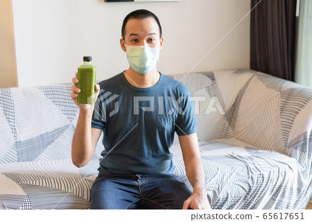 Young Asian man with mask showing green smoothie for healthy eating at home under quarantine Young Asian man with mask showing green smoothie for healthy eating at home under quarantine 65617651