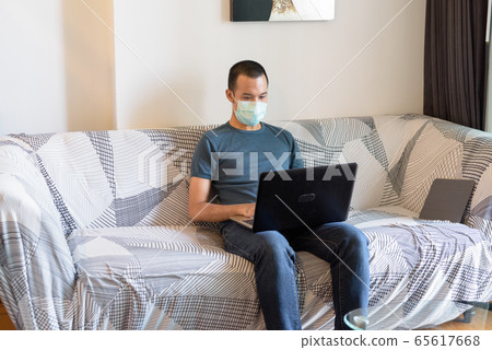 Young Asian man with mask using laptop at home under quarantine 65617668