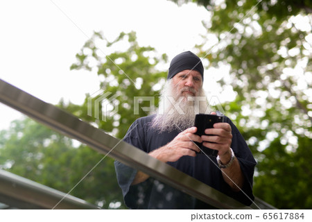 Mature bearded hipster man looking serious while using phone outside the subway train station Mature bearded hipster man looking serious while using phone outside the subway train station 65617884