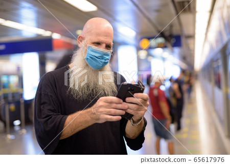 Mature bald bearded man with mask using phone at the subway train station Mature bald bearded man with mask using phone at the subway train station 65617976