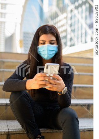 Young Indian woman with mask using phone and sitting by the stairs in city 65618903