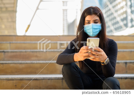 Young Indian woman with mask using phone and sitting by the stairs in city Young Indian woman with mask using phone and sitting by the stairs in city 65618905