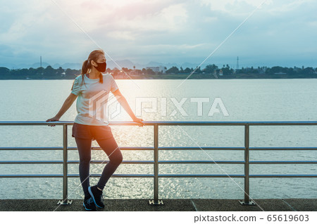 Woman standing resting tired with wearing a protective mask COVID-19 after running and jogging an outdoor workout on the riverwalk in the morning for lifestyle health. Woman standing resting tired with wearing a protective mask COVID-19 after running and jogging an outdoor workout on the riverwalk in the morning for lifestyle health. 65619603